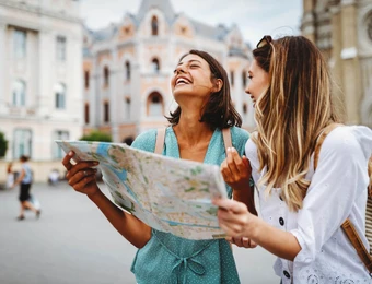 Zwei Frauen halten eine Karte; eine lacht, im Hintergrund Gebäude in einer Stadt.