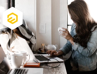 Eine Frau sitzt mit Laptop am Tisch und hält ein Smartphone. Links ein weißes Hexagon-Logo.