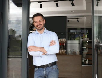 Mann in hellblauem Hemd und Jeans steht mit verschränkten Armen in offener Glastür eines Innenraums und lächelt.