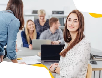 Eine Frau sitzt im Vordergrund am Tisch, lächelt in die Kamera; im Hintergrund arbeiten mehrere Personen mit Laptops.