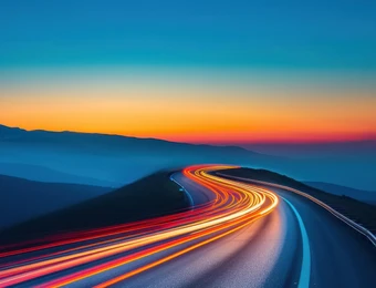 Kurvige Straße mit Lichtspuren, Berglandschaft im Hintergrund, Sonnenuntergang mit blauem und orangefarbenem Himmel.
