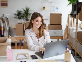 Eine Frau sitzt an einem Schreibtisch mit Laptop, umgeben von Kartons, einem Kleiderständer und Büro-Accessoires.