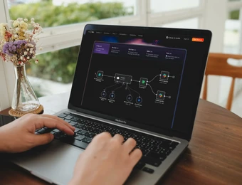Person tippt auf Laptop; Bildschirm zeigt Diagramm/Flowchart. Vase mit Blumen links vom Laptop; Fenster im Hintergrund.