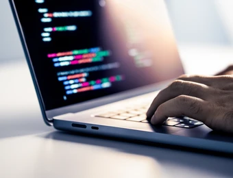 Nahaufnahme eines Laptops mit farbigem Code im Display; eine Hand tippt auf der Tastatur.