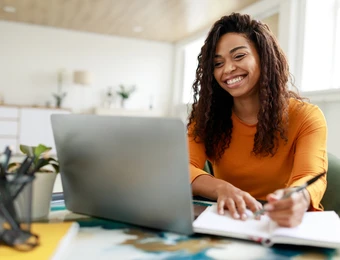 Eine Frau mit lockigen Haaren lächelt, sitzt an einem Schreibtisch vor dem Laptop und offenem Notizblock.