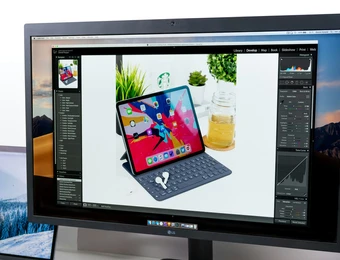 Monitor zeigt Fotobearbeitung, darauf ein iPad mit Tastaturhülle und Stift auf weißem Tisch, Pflanze im Hintergrund.