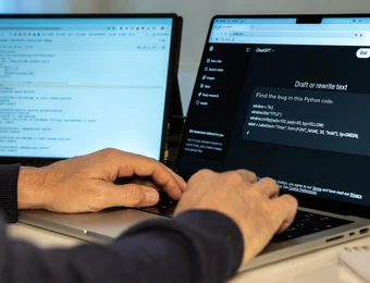Person tippt auf einem Laptop; zwei Monitore im Hintergrund zeigen Code und ein Chatfenster.
