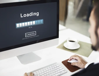 Computerbildschirm zeigt Ladebalken 'Loading' ca. 70% und 'CANCEL'; Tastatur, Maus, Kaffeetasse; Person am Schreibtisch.