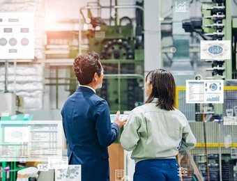 Zwei Personen in Business-Outfits stehen in einer Industrieanlage vor Monitoren, Diagrammen und Maschinen.