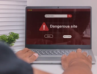 Laptop auf weißem Tisch; Bildschirm zeigt Warnung Dangerous site mit rotem Dreieck, Hände auf der Tastatur.