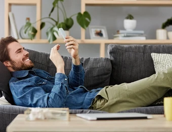 Mann liegt entspannt auf grauem Sofa, schaut auf Smartphone, trägt Jeansjacke und grüne Hose; Wohnzimmer mit Pflanzen.