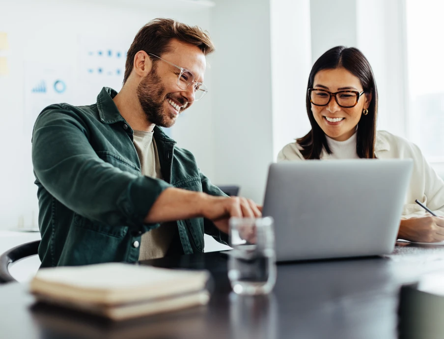 Zwei lächelnde Personen sitzen an einem Tisch mit Laptop; Wasser, Notizbuch, helles Büro im Hintergrund.