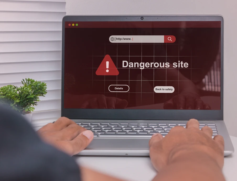Laptop auf weißem Tisch; Bildschirm zeigt Warnung Dangerous site mit rotem Dreieck, Hände auf der Tastatur.