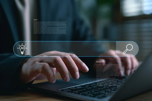 Nahaufnahme: Person tippt auf Laptop; holografische Suchleiste mit AI-Icon links, Lupe rechts.