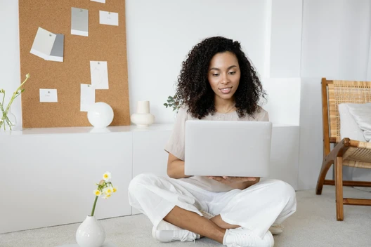 Person mit lockigen Haaren sitzt im hellen Wohnzimmer auf dem Boden und arbeitet an einem Laptop.