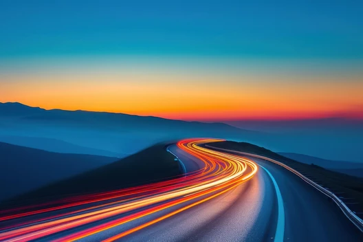 Kurvige Straße mit Lichtspuren, Berglandschaft im Hintergrund, Sonnenuntergang mit blauem und orangefarbenem Himmel.