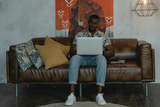 Mann sitzt auf braunem Ledersofa, Laptop im Schoß; Wand mit orangefarbener Kunst, mehrere Kissen.