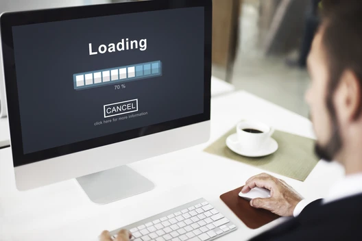 Computerbildschirm zeigt Ladebalken 'Loading' ca. 70% und 'CANCEL'; Tastatur, Maus, Kaffeetasse; Person am Schreibtisch.
